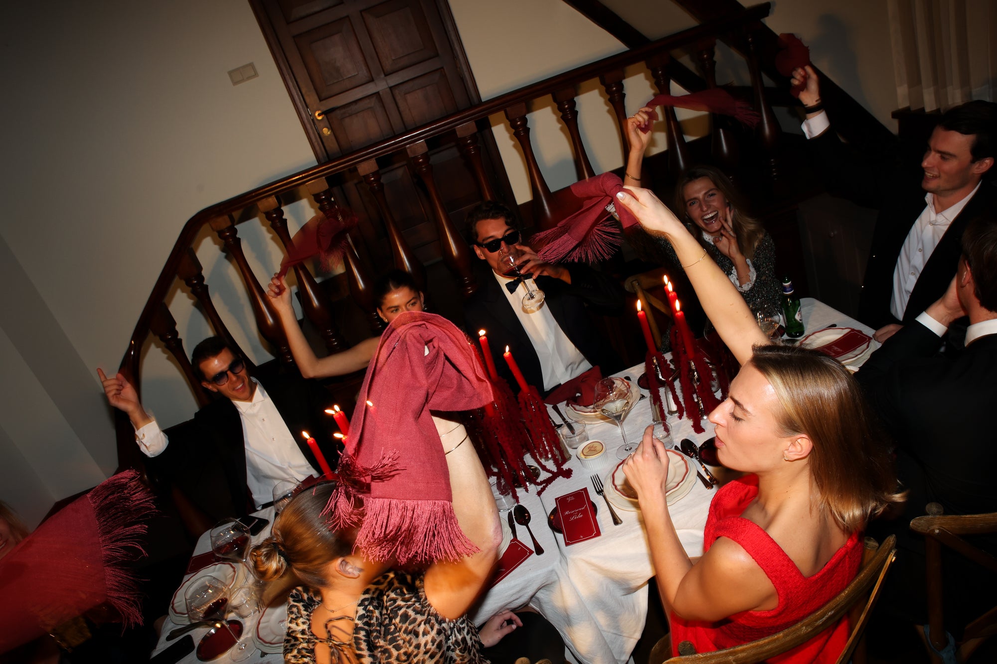 Guests celebrating around a vibrant dinner table styled by Imaginary Friends with red linen, glassware, and layered plates.