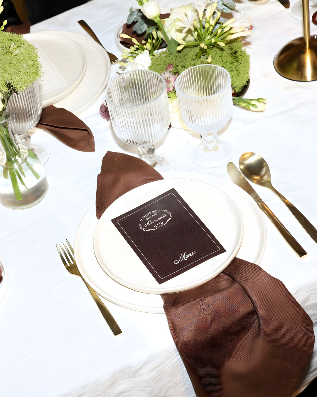 Close-up of The Suite table styling with personalized brown menu, white plate set, brown linen napkin, gold cutlery and decorative florals by Imaginary Friends.