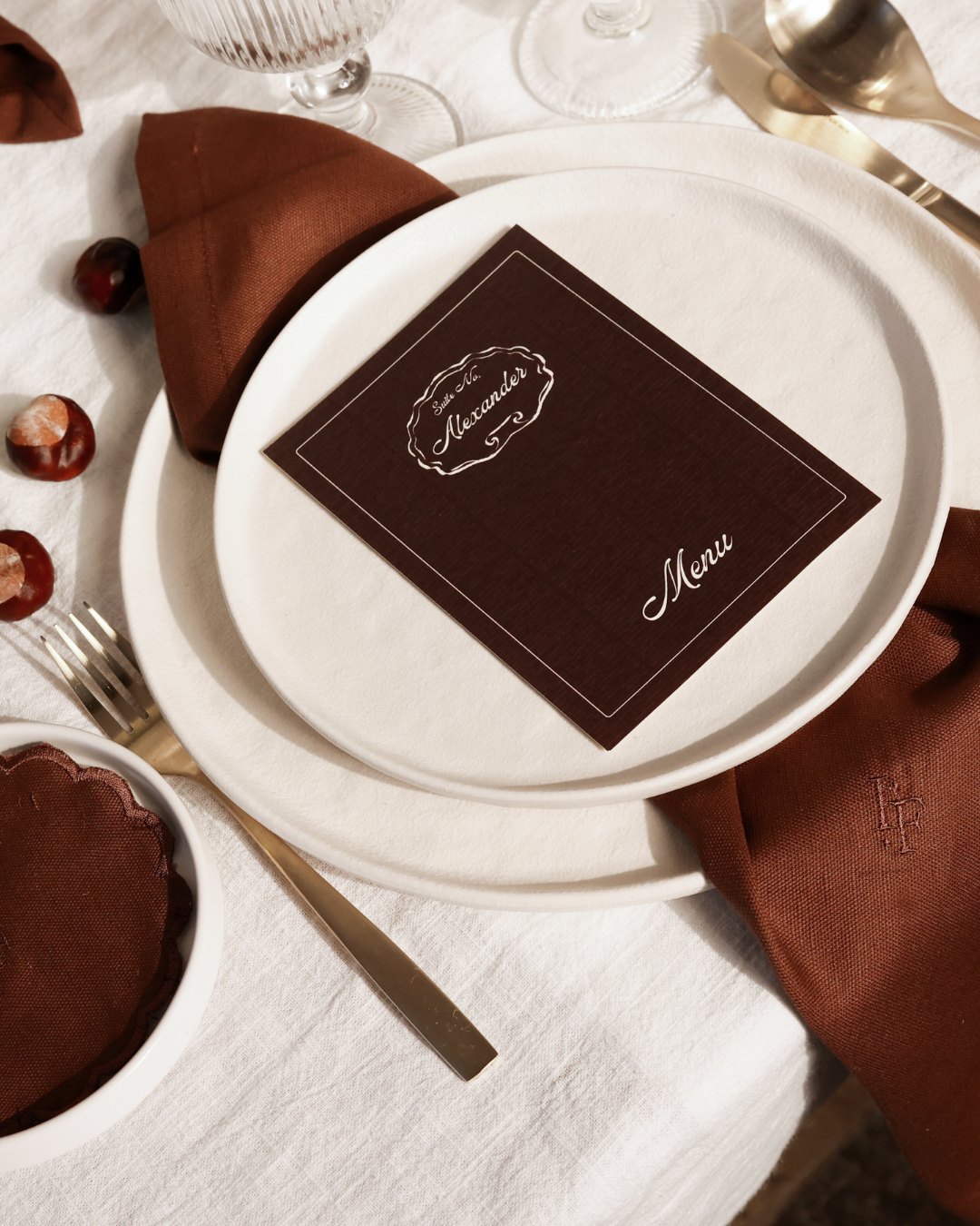 Close-up of The Suite table styling by Imaginary Friends with layered white plates, brown linen napkin, personalized brown menu card and gold cutlery.