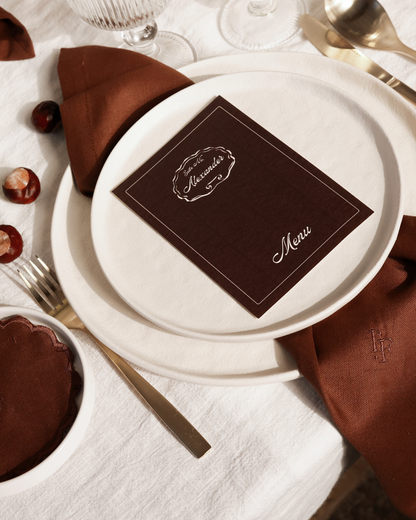 Close-up of The Suite table styling by Imaginary Friends with layered white plates, brown linen napkin, personalized brown menu card and gold cutlery.