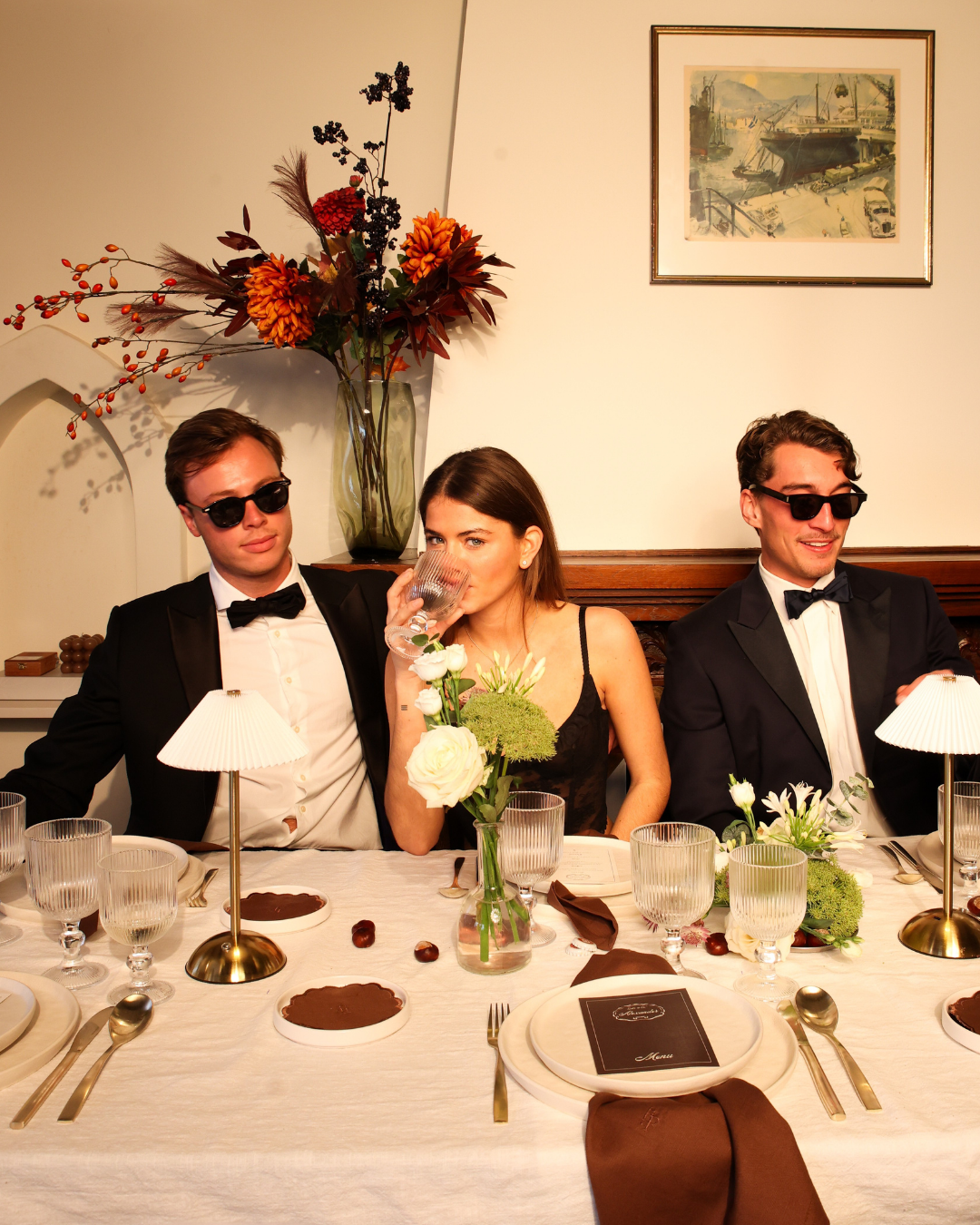 Three guests in formal attire seated at The Suite dinner table by Imaginary Friends, with gold accents, ribbed glassware, white table linen and seasonal florals.