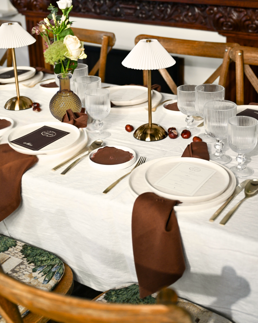 Full The Suite tablescape by Imaginary Friends featuring white tablecloth, gold table lamps, ribbed clear glasses, brown linen napkins and seasonal floral arrangements.