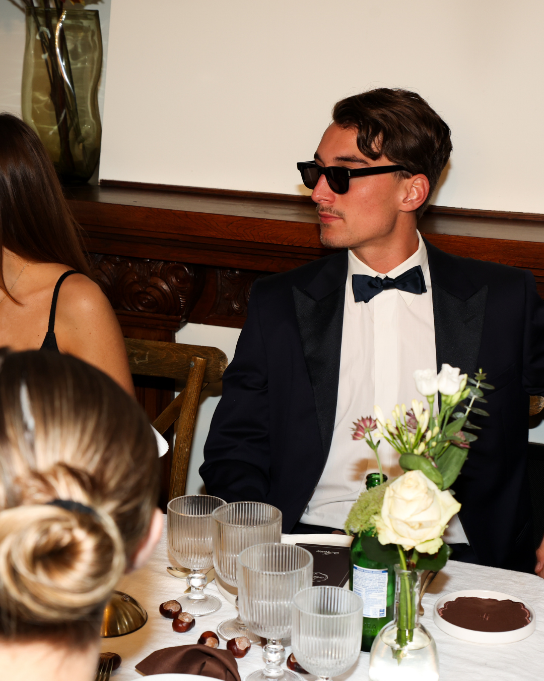 Elegant dinner setting from The Suite by Imaginary Friends, featuring a male guest in a tuxedo seated at a styled table with white plates, ribbed glassware and neutral florals.